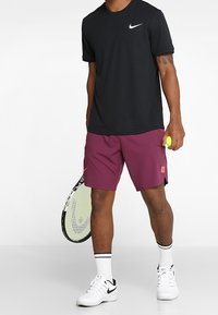 Muž v černém tričku Nike, vínových šortkách a bílých teniskách Nike drží tenisovou raketu a žlutý tenisový míček před jednobarevným pozadím.