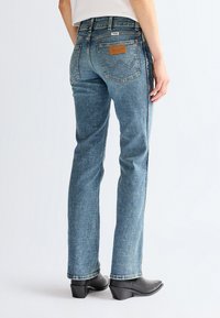Person trägt mittelblaue Wrangler Straight-Leg-Jeans und schwarze Stiefeletten mit Absatz, von hinten vor einfarbigem Hintergrund gezeigt.
