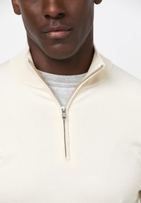 Cremefarbener Pullover mit einem Halb-Reißverschluss-Kragen und gerippten Akzenten, über ein hellgraues Hemd getragen. Weicher, strukturierter Stoff.