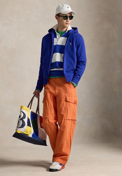 Homme portant une casquette blanche, des lunettes de soleil, un sweat à capuche bleu zippé, une chemise rayée, un pantalon cargo orange, des baskets blanches, tenant un sac fourre-tout coloré.