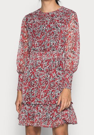 Kobieta ubrana w długą sukienkę z czerwonym kwiatowym wzorem, z marszczonym pasem, mankietami na rękawach i falbanami na dole.