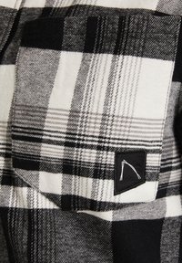 Zwart-witte geruite flanellen stof met een gestructureerd patroon; heeft een vierkante zak en een zwart etiket met een wit logo.