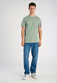 Młody mężczyzna stoi, ubrany w zieloną koszulkę Lindbergh, niebieskie dżinsy i białe trampki, na tle jednolitego, jasnego tła.