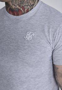 Camiseta corta de manga gris en una tela suave y texturizada. Presenta un logo bordado en blanco en el lado izquierdo del pecho. Cuello redondo.