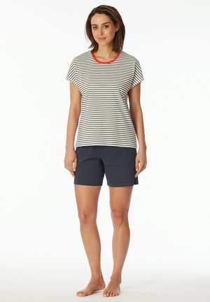 Frau steht barfuß in einem weiß und schwarz gestreiften T-Shirt mit rotem Kragen und dunkelblauen Shorts vor einem weißen Hintergrund.