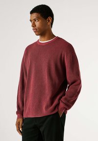 Roter Pullover mit strukturierter Oberfläche, langen Ärmeln und rundem Ausschnitt, kombiniert mit dunkler Hose. Der Stoff weist ein geripptes Muster auf.