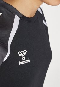 Prikaz izbliza osobe koja nosi crnu sportsku majicu s bijelim prugama na ramenima i bijelim Hummel logom na prsima.