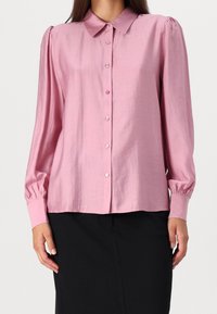 Chemise rose à manches longues avec boutons et col, dotée d'une texture lisse et de manches bouffantes, associée à un bas noir.