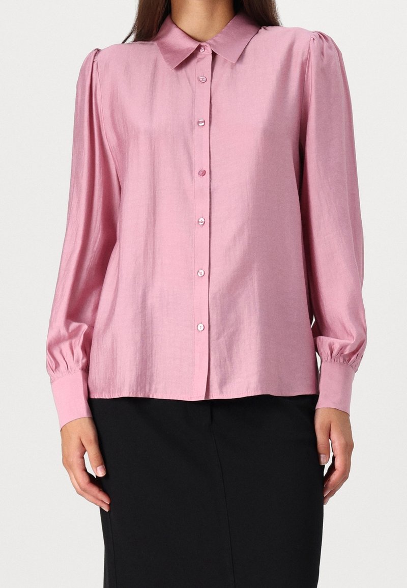 Chemise rose à manches longues avec boutons et col, dotée d'une texture lisse et de manches bouffantes, associée à un bas noir.