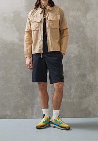 Giacca leggera beige con due tasche sul petto, t-shirt nera, pantaloni cargo neri, sneakers gialle e blu con calzini bianchi, sfondo grigio testurizzato.