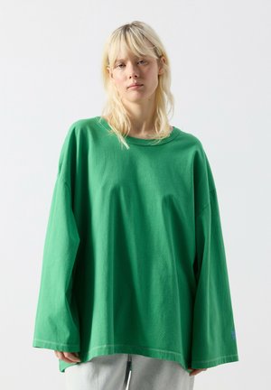 Šviesiaplaukis asmuo dėvi per didelį žalią ilgomis rankovėmis marškinius ir šviesiai pilką kelnes, stovintis prieš paprastą šviesią foną.