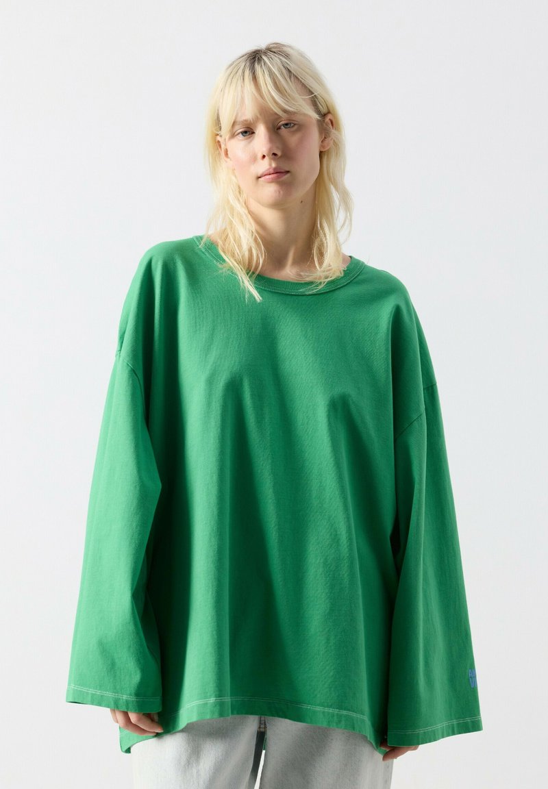 Blond osoba w oversize'owej zielonej koszuli z długim rękawem i jasnoszarych spodniach, stojąca na tle gładkiego, jasnego tła.