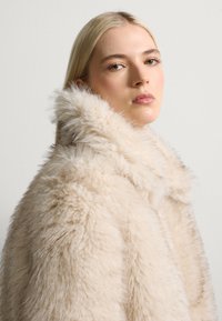 Cappotto in pelliccia sintetica di un beige chiaro con una consistenza spessa e morbida; presenta un colletto alto e una silhouette slanciata.