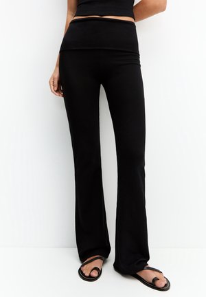 Femme portant un pantalon noir taille haute évasé avec un haut court noir et des sandales noires à bout ouvert sur un fond blanc.