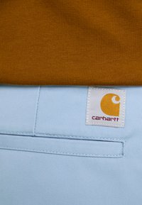 Carhartt genser i sennepsfarge over lyseblå bukser, med et hvitt merke med en gul logo og rød tekst. Strukturert strikking og sømmer synlige.