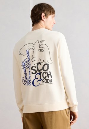 Mężczyzna stojący bokiem, ubrany w kremową bluzę z abstrakcyjną grafiką ptaka i twarzy oraz napisami „Dreamers Gonna Dream” i „Scotch & Soda” na plecach.