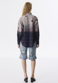 Overmaatse geblokte blouse in tinten paars en blauw, met versleten accenten, gecombineerd met gefronde blauwe denim shorts en zwarte hakken.