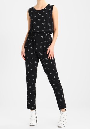 Woman wearing a sleeveless black jumpsuit with white bird patterns, paired with white ankle boots with grommet details.