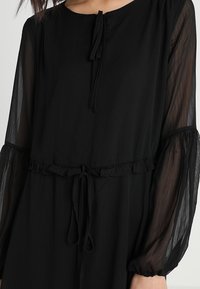 Schwarzes Kleid mit langen, durchsichtigen Ärmeln, rundem Ausschnitt, gerafftem Taillenbund und gebundenem Detail an der Vorderseite. Leichter, strukturierter Stoff.