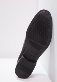 Semelle de chaussure noire avec une surface texturée et côtelée. Comprend une incrustation rectangulaire et un bord incurvé, offrant adhérence et durabilité.