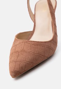 Καφέ σουέντ γόβα με μυτερή μύτη και λουράκι πίσω, με υφή τύπου κροκό και μπεζ ενισχυμένο πάτο, σε λευκό φόντο.