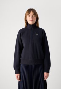Young woman wearing a navy blue quarter-zip sweatshirt and matching pleated skirt, standing against a plain white background.
