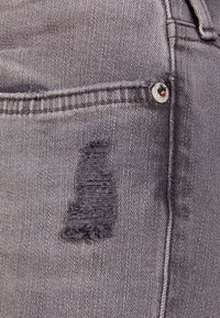 Grå denimtyg har ett slitet område med fransade kanter. Designen inkluderar en metallknapp med kopparaccent och standardstygn.