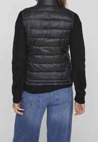 Svart pufferväst med en figurnära design och horisontellt quiltad textur, bärs över en kraftig svart stickad tröja och matchas med blå vida jeans.