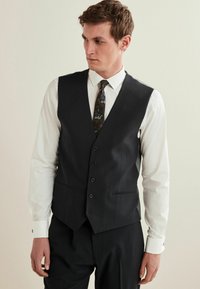 Schwarze Weste mit maßgeschneidertem Design, fünf Knöpfen und Seitentaschen, kombiniert mit einem weißen Hemd und einer gemusterten Krawatte. Glatte Stofftextur.