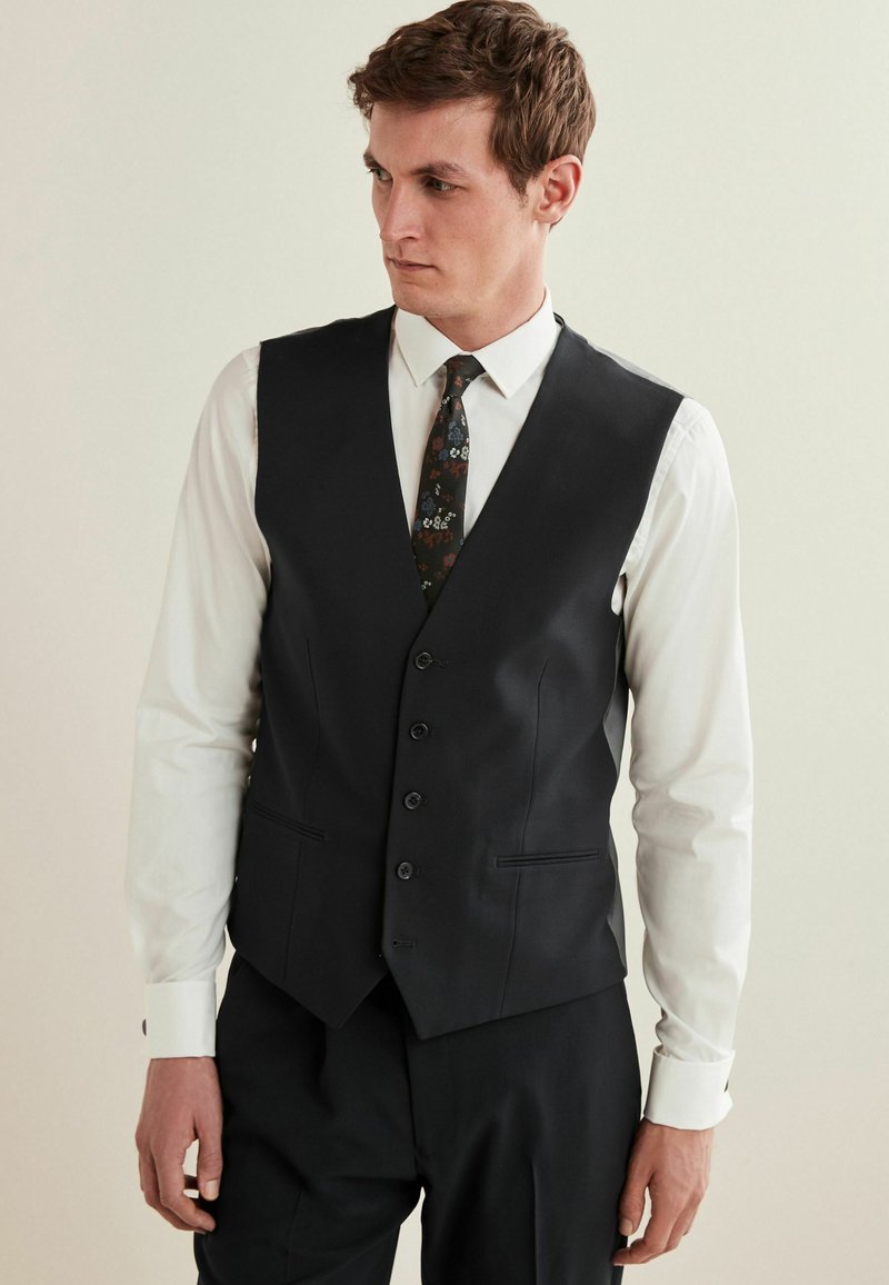 Schwarze Weste mit maßgeschneidertem Design, fünf Knöpfen und Seitentaschen, kombiniert mit einem weißen Hemd und einer gemusterten Krawatte. Glatte Stofftextur.