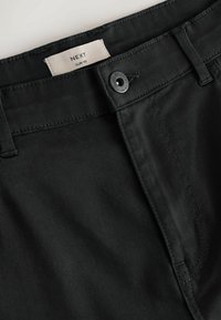 Pantalon noir slim avec fermeture par bouton et passants pour ceinture, doté d'une étiquette blanche avec le nom de la marque « NEXT » à l'intérieur de la ceinture.