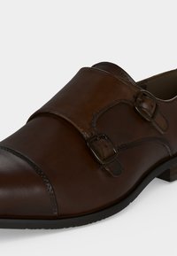Chaussure habillée en cuir marron avec boucles à double bride moine et semelle noire sur fond blanc.