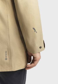 Beige Gore-Tex-Jacke mit glatter Textur, taillierten Ärmeln und einem dezenten Logo-Detail nahe dem Bündchen. Knopfverschluss am Handgelenk.