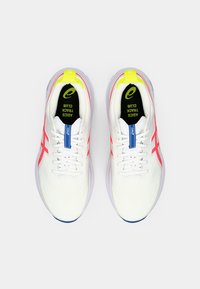 White athletic shoes with a breathable upper, pink accents, a yellow heel tab, and a blue lace lock. Features a textured midsole and rubber outsole.