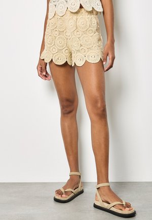 Beige gehäkelte Shorts mit kreisförmigen Mustern und gewellten Kanten, getragen mit beigen flachen Sandalen mit Knöchelriemen.