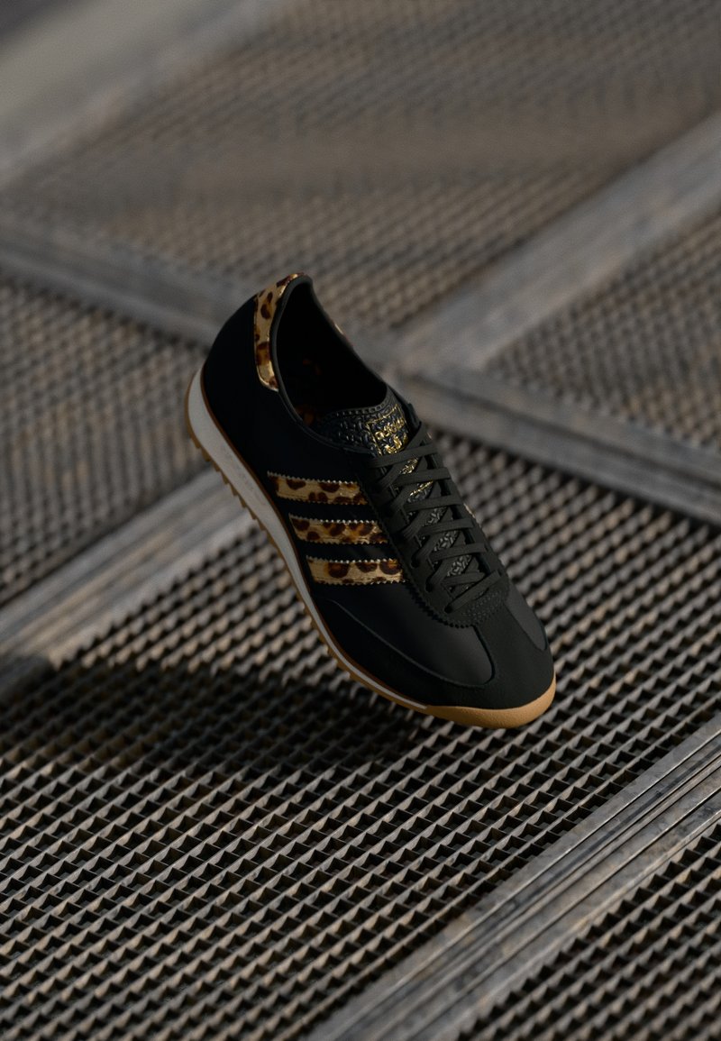 Black athletic shoe with leopard print accents, textured toe box, smooth leather, and three gold stripes. Lightweight rubber sole.
