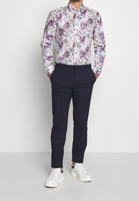 Camicia a fiori con accenti viola, verde e rosa; pantaloni sartoriali blu navy e sneaker bianche. Design slim fit, colletto con bottoni.
