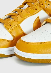 Close-up of white and mustard yellow leather sneakers with perforated toe box and woven laces on white background.