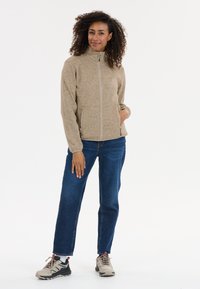 Ljust brunt fleecejacka med hög krage och blixtlås, utrustad med två sidofickor, bärs med blå jeans och beige och svarta sneakers.