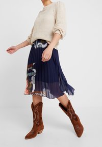 Beige pletený svetr s nabíranými rukávy, kombinovaný s námořnickou skládací sukní s květinovými vzory a hnědými semišovými kovbojskými botami s vyšíváním.