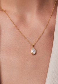 Złoty łańcuszek naszyjnika z okrągłym, przezroczystym zawieszką otoczonym mniejszymi przezroczystymi kamieniami, spoczywający na skórze.