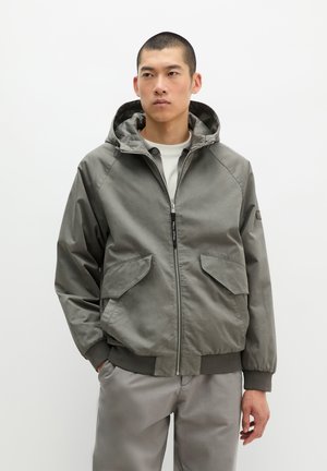 Mann trägt eine graue Kapuzenjacke mit Reißverschluss und großen Klappentaschen sowie graue Hose, steht vor einem schlichten weißen Hintergrund.