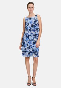 Niebieska sukienka w kwiaty z warstwowymi falbanami, wykonana z lekkiego materiału. Posiada okrągły dekolt i sięga do połowy uda. Czarne sandały na obcasie.