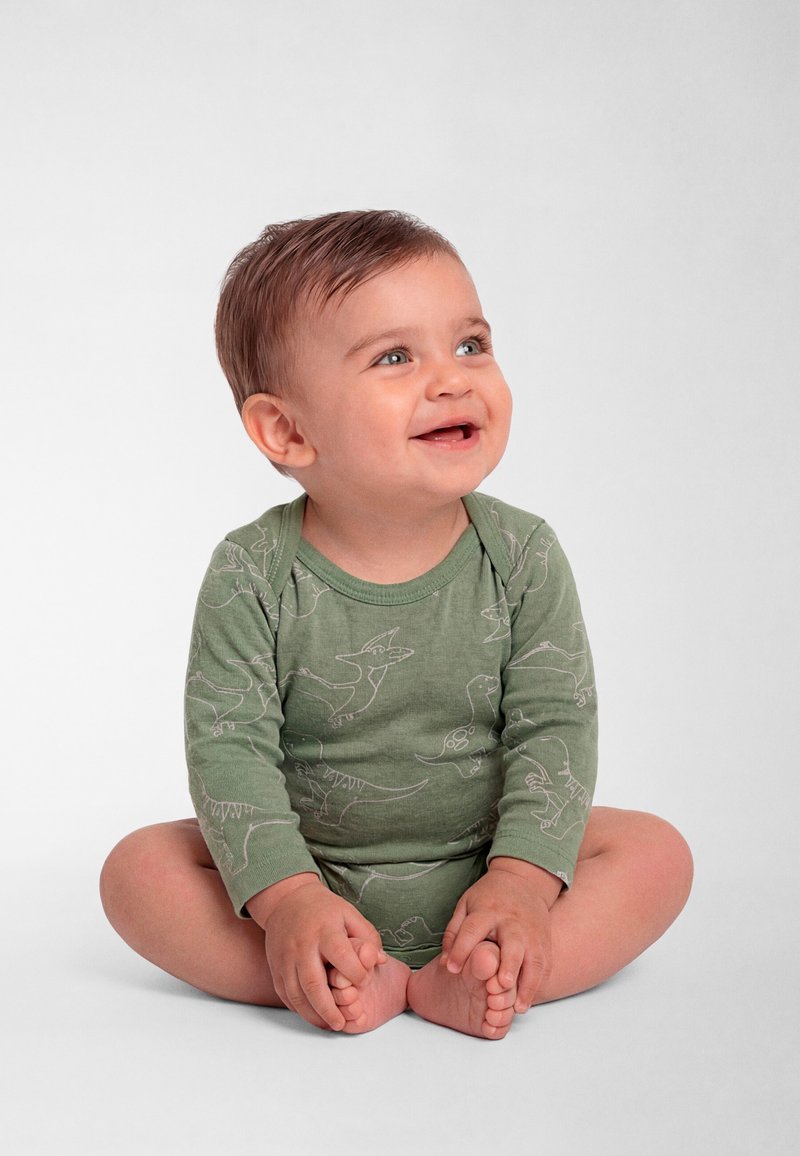 Zamišljeno dijete koje sjedi prekriženih nogu, nosi zelenu dugoručnu pidžamu s otiscima dinosaura na jednobojnoj bijeloj pozadini.