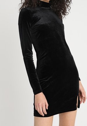 Kvinde iført en tætsiddende, langærmet, sort velvet minikjole med høj hals og en lille sidespring.