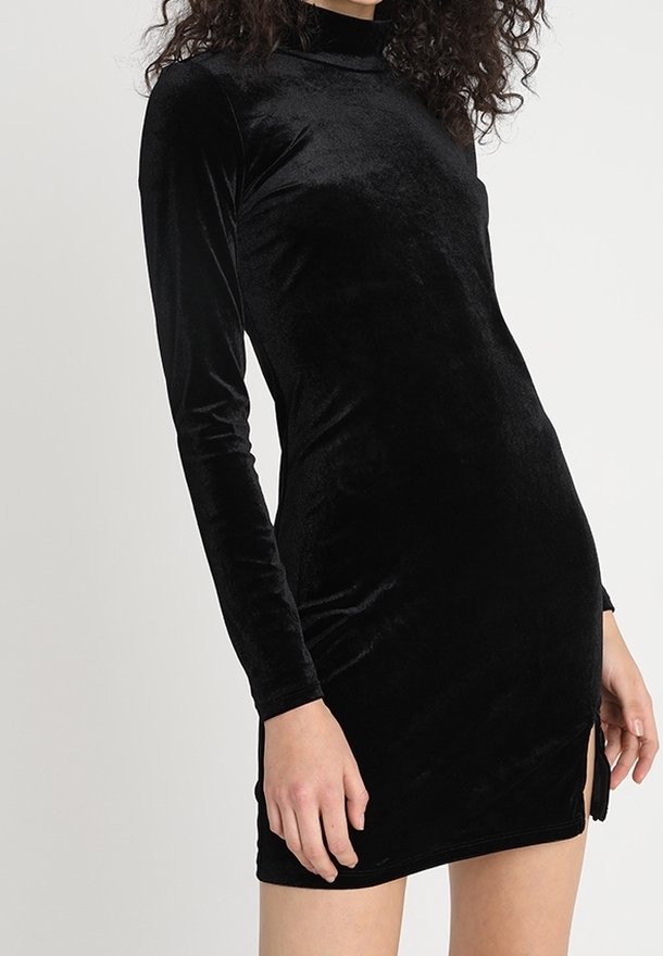 Femme portant une mini-robe moulante en velours noir à manches longues, avec un col haut et une petite fente latérale.