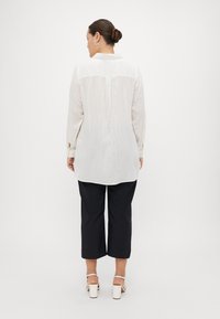Chemise blanche à manches longues avec des rayures verticales subtiles et des détails de boutons, associée à un pantalon noir cropped, sur fond neutre.