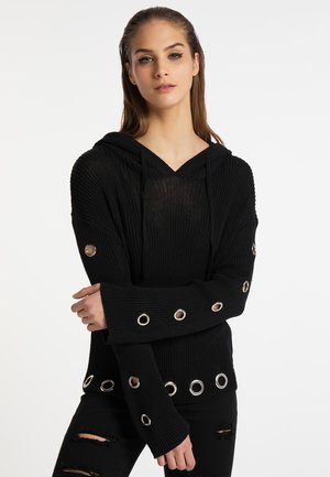 Jonge vrouw draagt een zwarte gebreide hoodie met metalen oogjes op de mouwen en zoom, gecombineerd met zwarte gescheurde jeans, poseert met gekruiste armen.