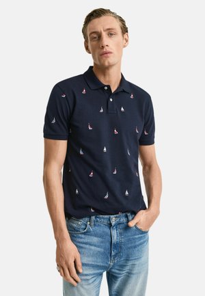 Marineblaues Poloshirt mit Segelboot-Stickerei in Rot und Weiß, kurzen Ärmeln und klassischem Kragen, kombiniert mit hellblauen Jeans.