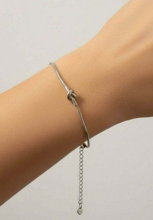 Zilveren armband met een knoopontwerp, voorzien van een slanke ketting en een klein hartvormig bedel, gedragen om een pols met een huidskleur.